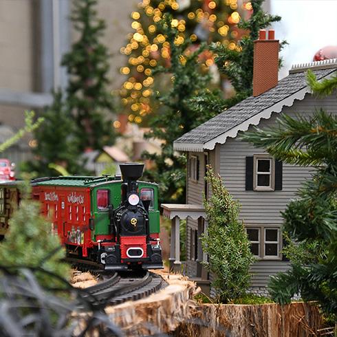 Miniature train running by model building with lit Christmas tree in the background